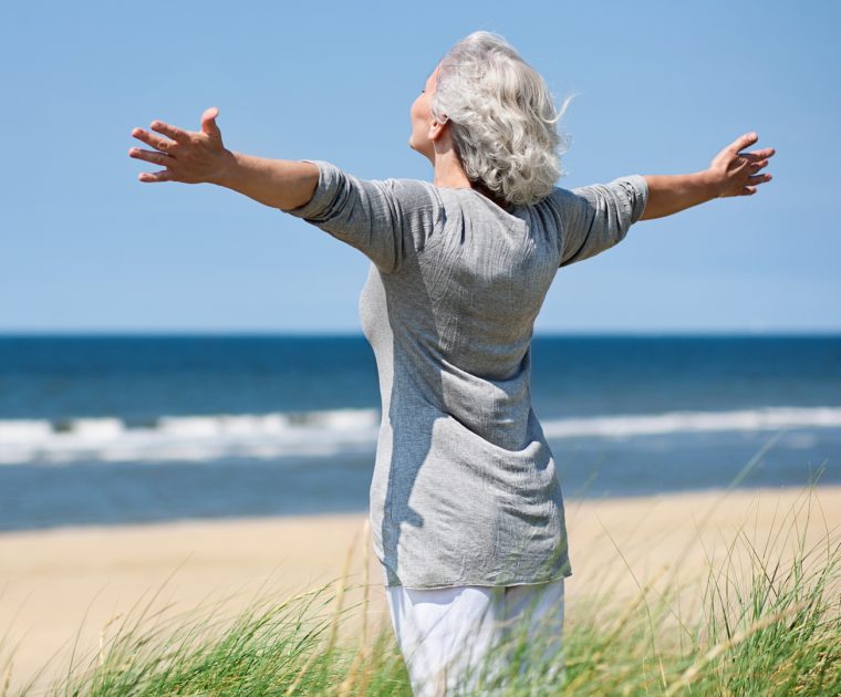 An older lady with grey hair standing with arms outstretched on a beach