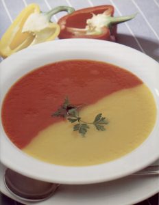 A white bowl containing two separate differently coloured soups. One half is a red pepper soup and the other half is a yellow pepper soup with no mix or overlap.
