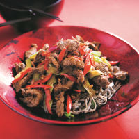A stirfry of Beef steak strips with ginger, sugar-snap peas, and pak choi on a bed of nnodles on a red oriental style bowl