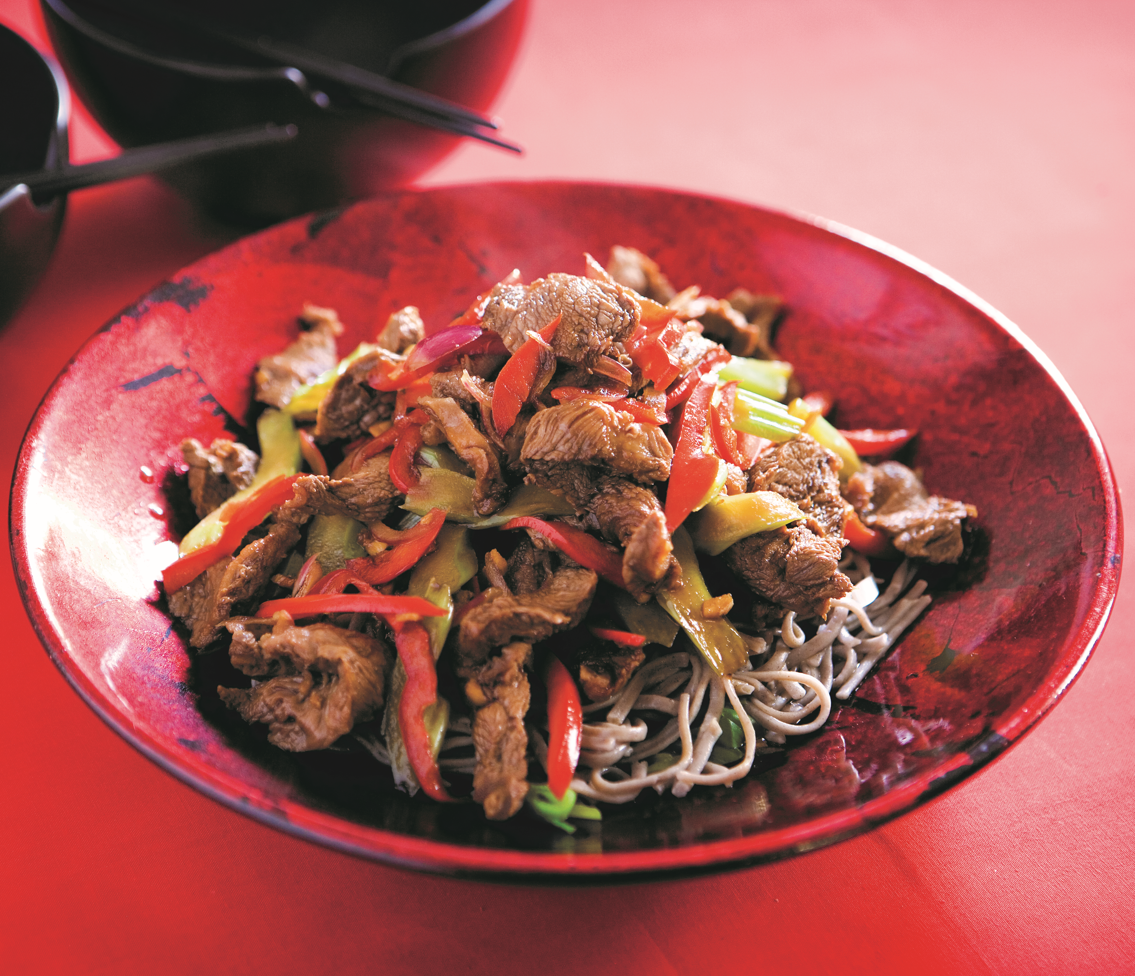 A stirfry of Beef steak strips with ginger, sugar-snap peas, and pak choi on a bed of nnodles on a red oriental style bowl