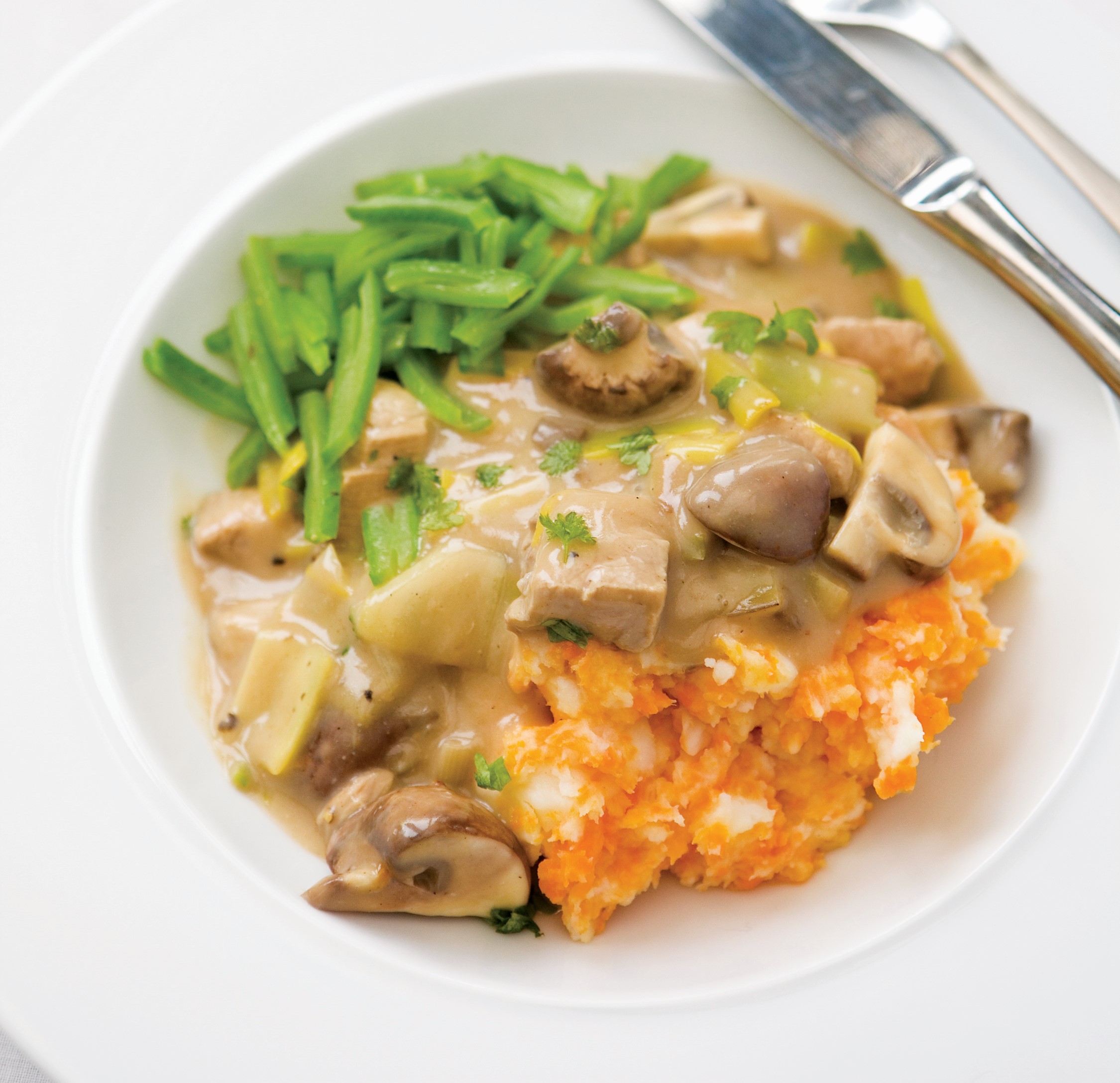 A creamy looking Pork and Leek Casserole on a white plate served with sweet potato mash and sliced green beans
