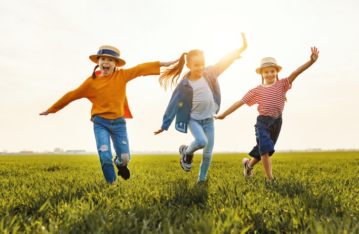 three children running across a grassy field in the sun looking like they are pretending to be aeroplanes