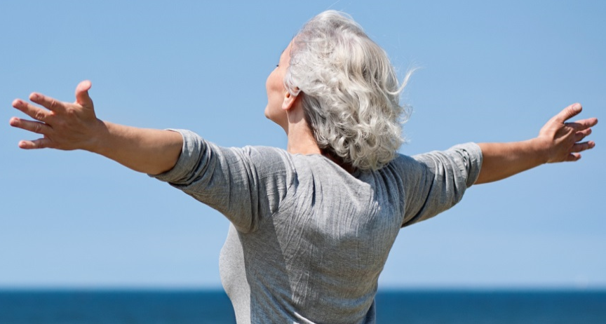 An older lady with grey hair standing with arms outstretched on a beach
