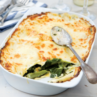 A white dish of Swiss Chard Lasagne with one portion scooped out showing the crisp golden layer of pasta on top with green chard layered with more pasta underneath in a creamy looking sauce