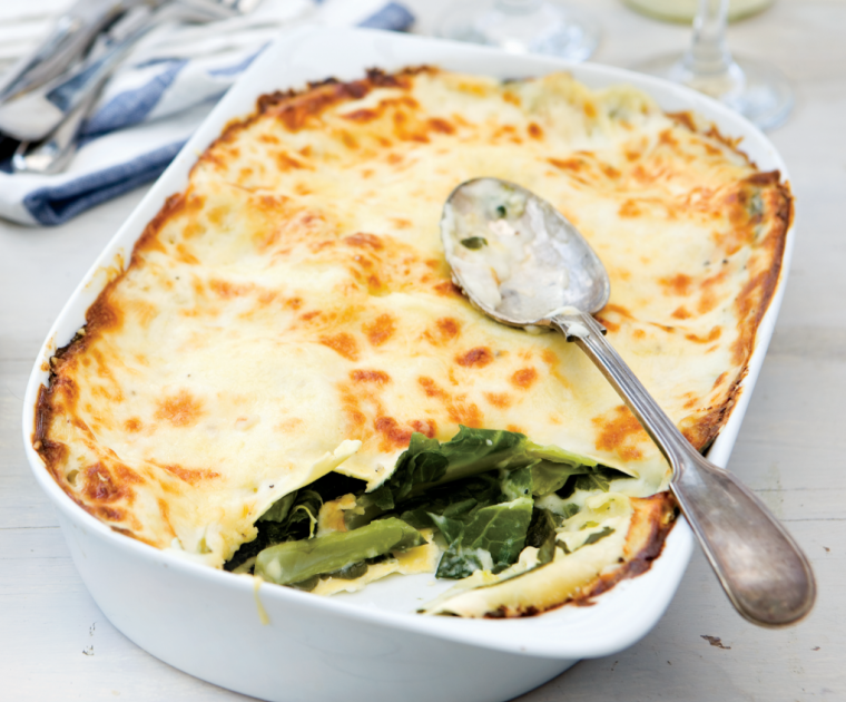 A white dish of Swiss Chard Lasagne with one portion scooped out showing the crisp golden layer of pasta on top with green chard layered with more pasta underneath in a creamy looking sauce