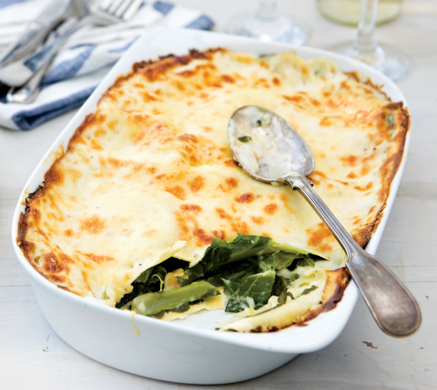 A white dish of Swiss Chard Lasagne with one portion scooped out showing the crisp golden layer of pasta on top with green chard layered with more pasta underneath in a creamy looking sauce