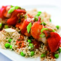A Chinese Chicken Kebab of chunks of chicken, red pepper and water chestnuts on a wooden skewer, marinaded in a chinese sauce resting on a bed of rice and peas.