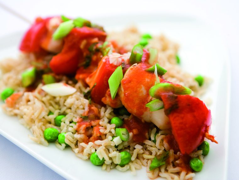 A Chinese Chicken Kebab of chunks of chicken, red pepper and water chestnuts on a wooden skewer, marinaded in a chinese sauce resting on a bed of rice and peas.