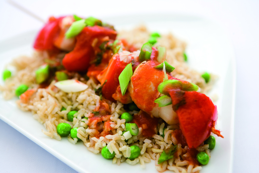 A Chinese Chicken Kebab of chunks of chicken, red pepper and water chestnuts on a wooden skewer, marinaded in a chinese sauce resting on a bed of rice and peas.