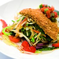A crispy breadcrumbed fillet of chicken resting on top of a plate of stir-fried vegetables.