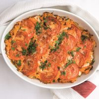 An oval pie dish containing Ham Leek & Sweet Potato Pie topped with slices of tomato and a grilled cheese topping