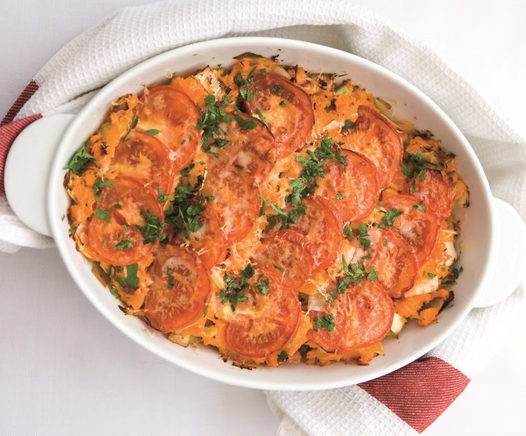 An oval pie dish containing Ham Leek & Sweet Potato Pie topped with slices of tomato and a grilled cheese topping