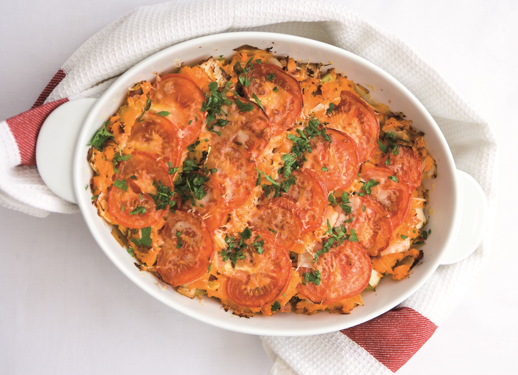 An oval pie dish containing Ham Leek & Sweet Potato Pie topped with slices of tomato and a grilled cheese topping