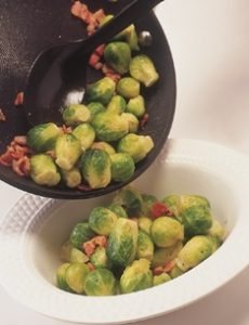 Tender green Brussells Sprouts mixed with crispy pancetta being poured out of a pan into a serving dish